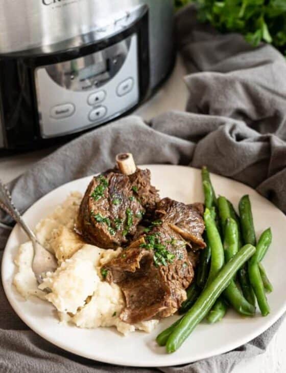Beef short ribs on a plate with mashed potatoes and green beans in front of a slow cooker.