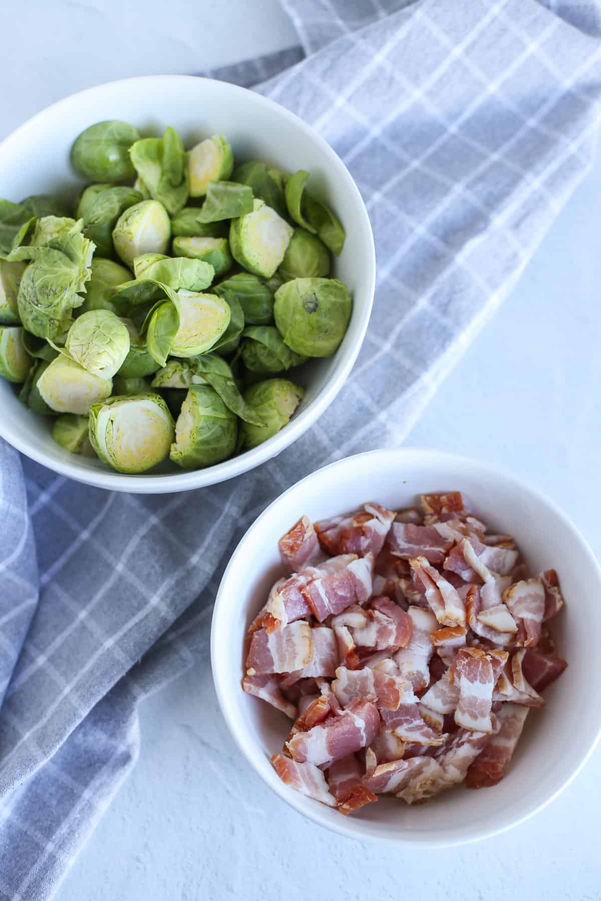 Bowl of brussels sprouts chopped in half and bowl of chopped bacon setting on a counter.