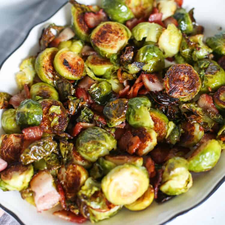 A hand holding a serving dish of roasted Brussels sprouts with bacon.