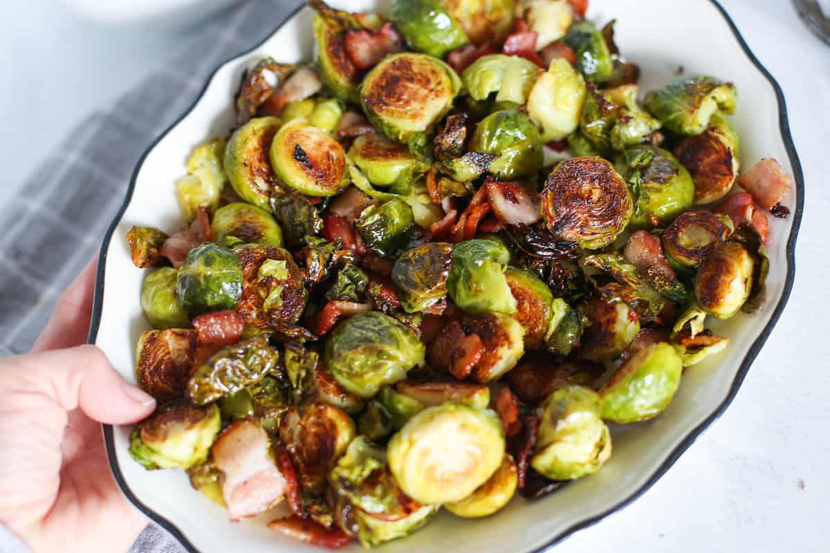 A hand holding a serving dish of roasted Brussels sprouts with bacon.