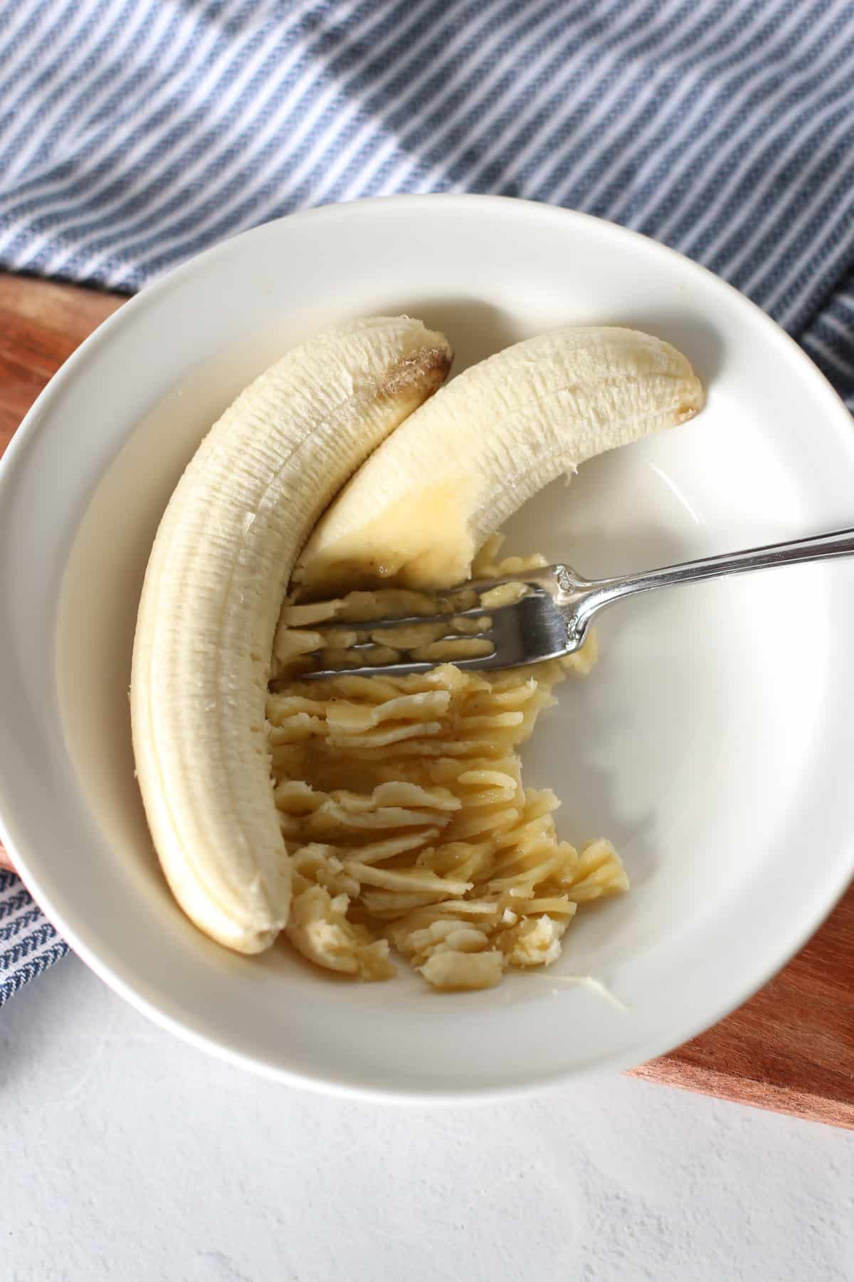 A fork mashing bananas in a white bowl.