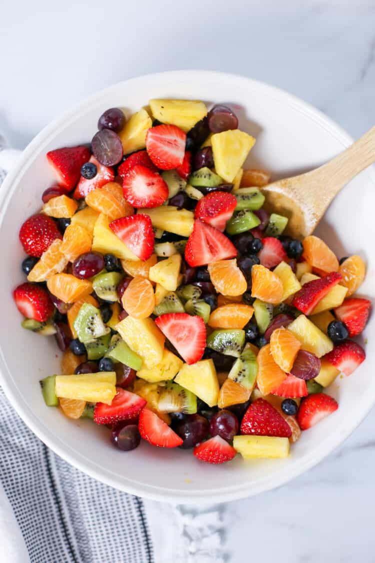 Fruit salad in a white serving bowl with a wooden spoon.