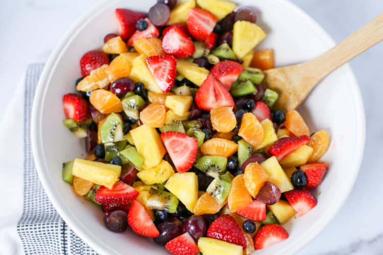 Fruit salad in a white bowl with a wooden spoon for serving.