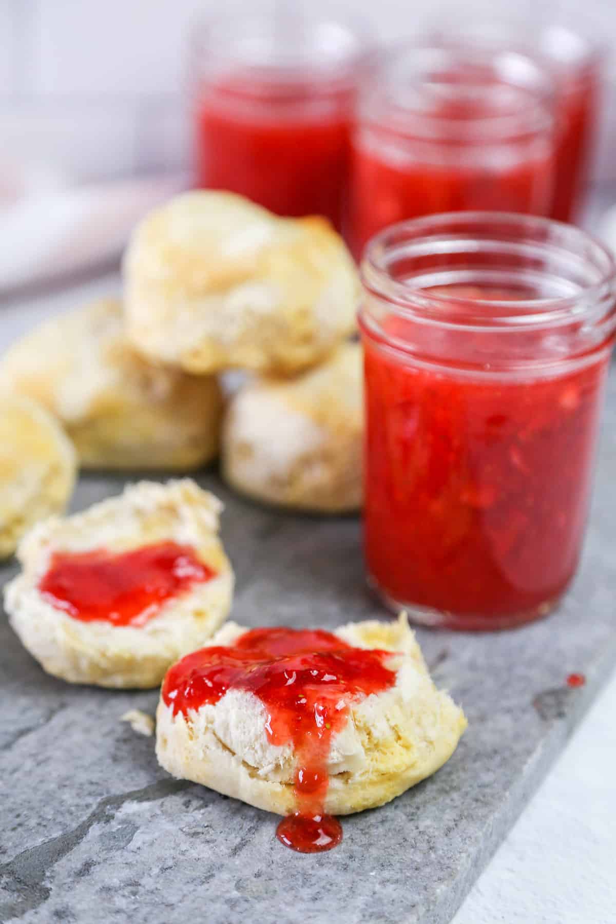 Strawberry freezer jam on biscuit halves with more biscuits and jelly jars stacked in the background.