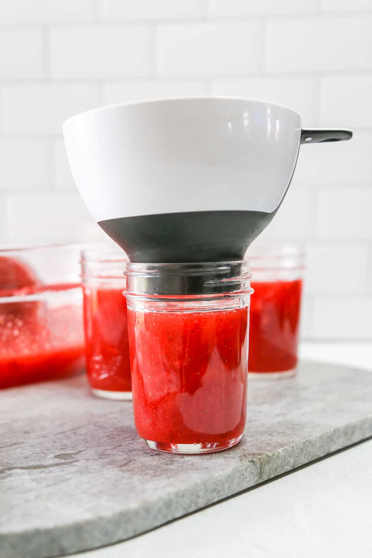 Adding strawberry jam to an 8-ounce mason jar using a funnel.