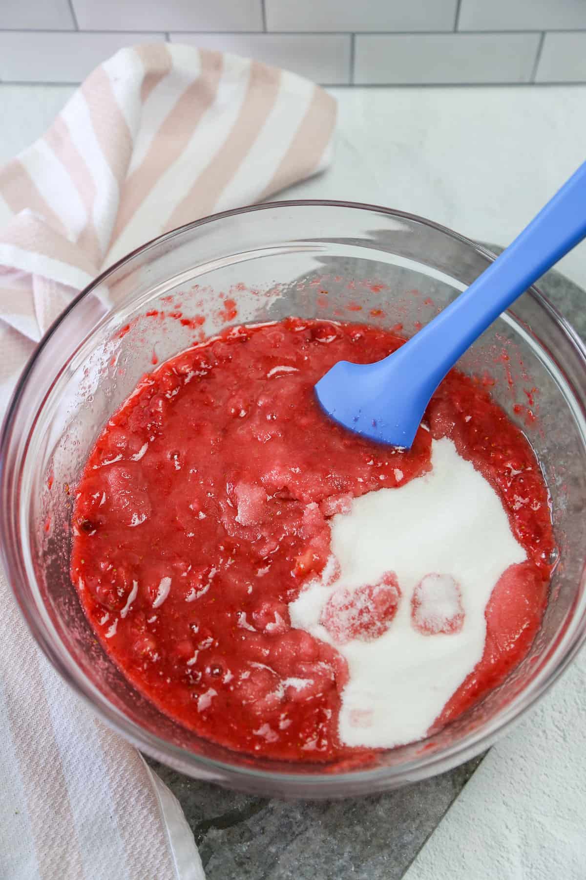 Sugar being mixed into crushed strawberries in a glass mixing bowl.