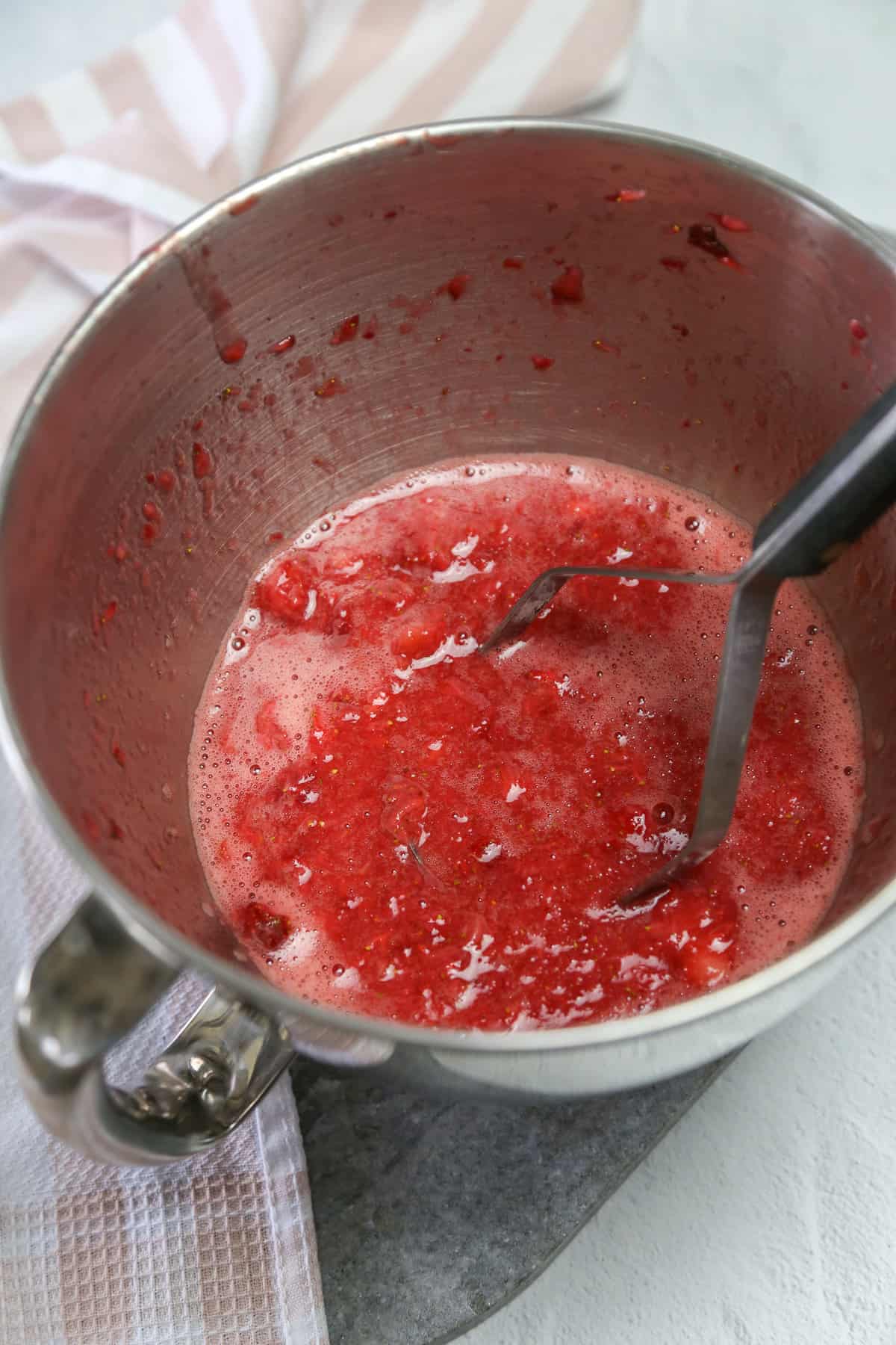 Potato masher crushing strawberries in a metal mixing bowl to make freezer jam.