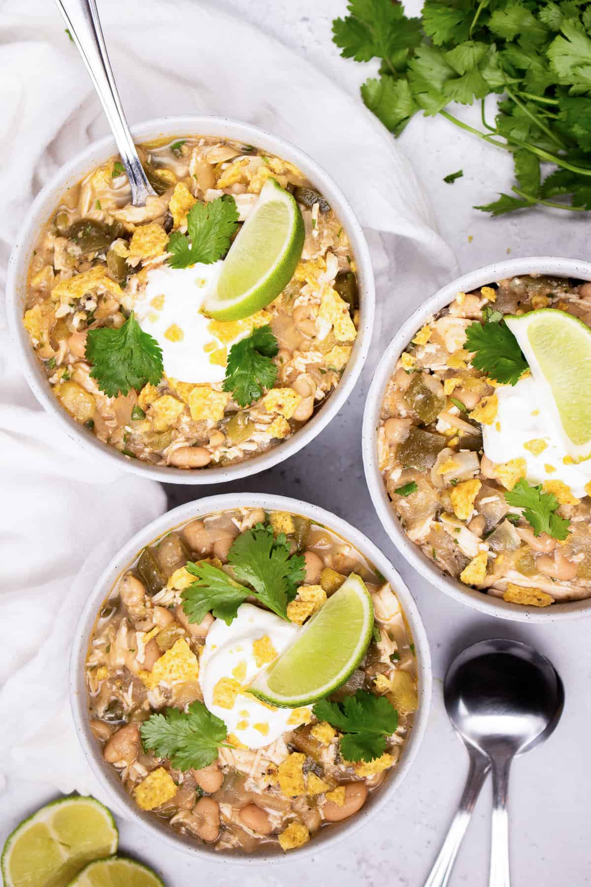 Several bowls of white chicken chili with sour cream, crushed tortilla chips, chopped cilantro, and a lime wedge on top.