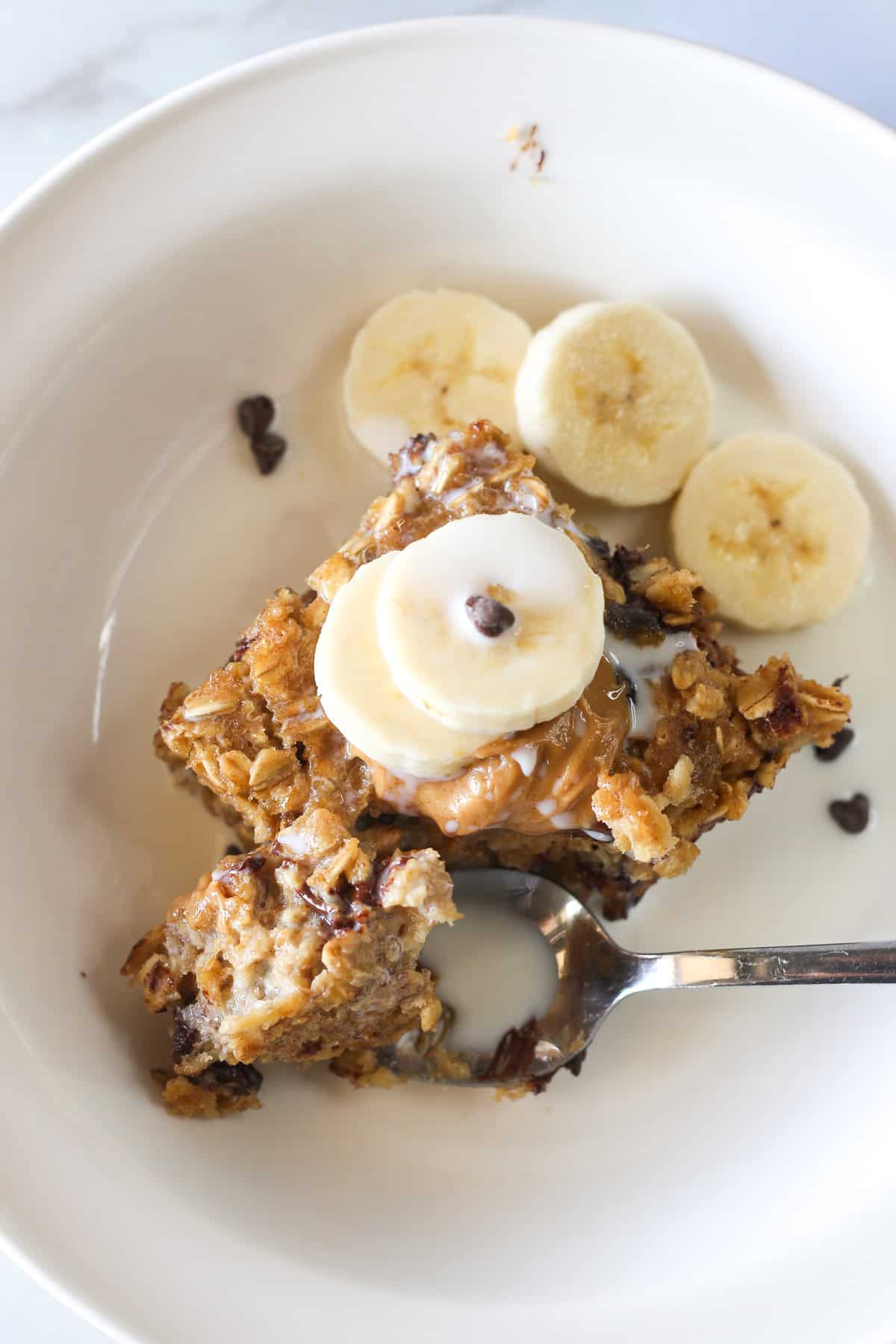 A slice of baked oatmeal in a bowl topped with milk, peanut butter, banana slices, and mini chocolate chips.