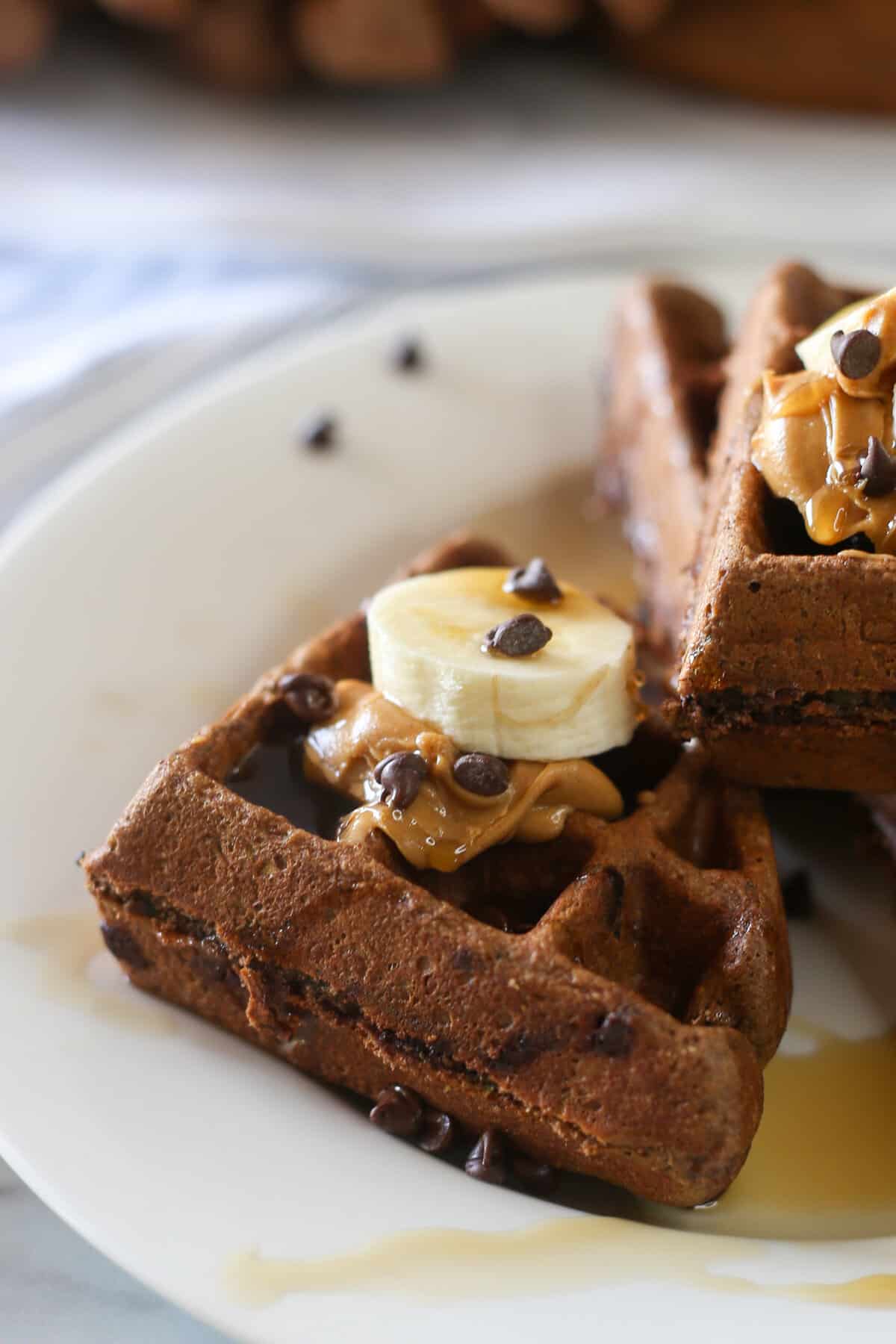 Chocolate zucchini waffles stacked on a plate with banana slices, peanut butter and mini chocolate chips on top.