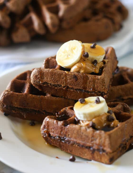 Chocolate zucchini waffles stacked on a plate and topped with banana slices, peanut butter, and mini chocolate chips.