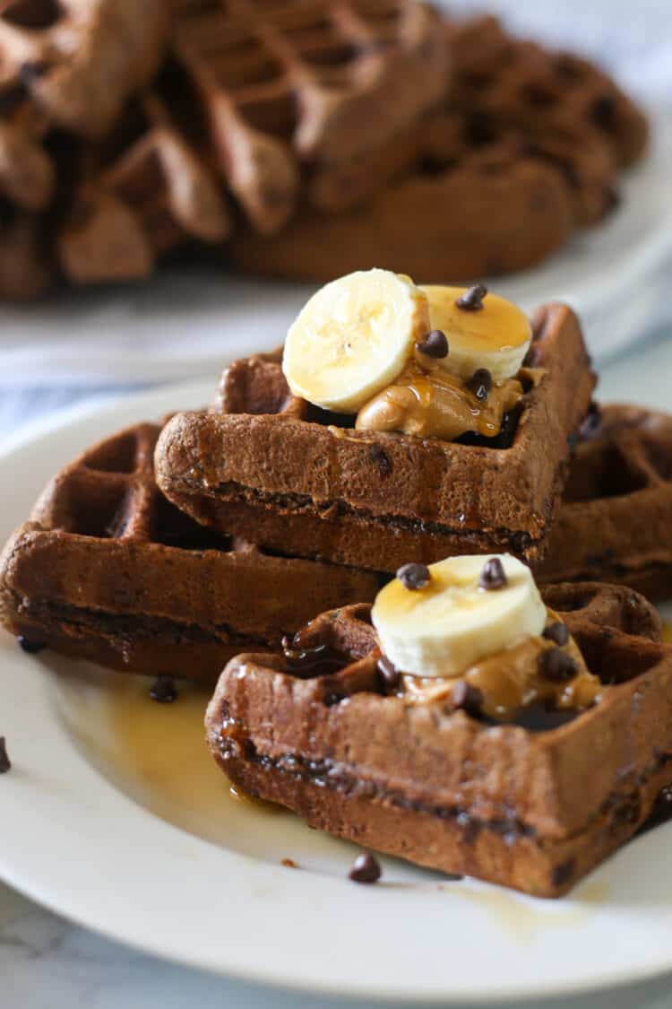 Chocolate zucchini waffles stacked on a plate and topped with banana slices, peanut butter, and mini chocolate chips.