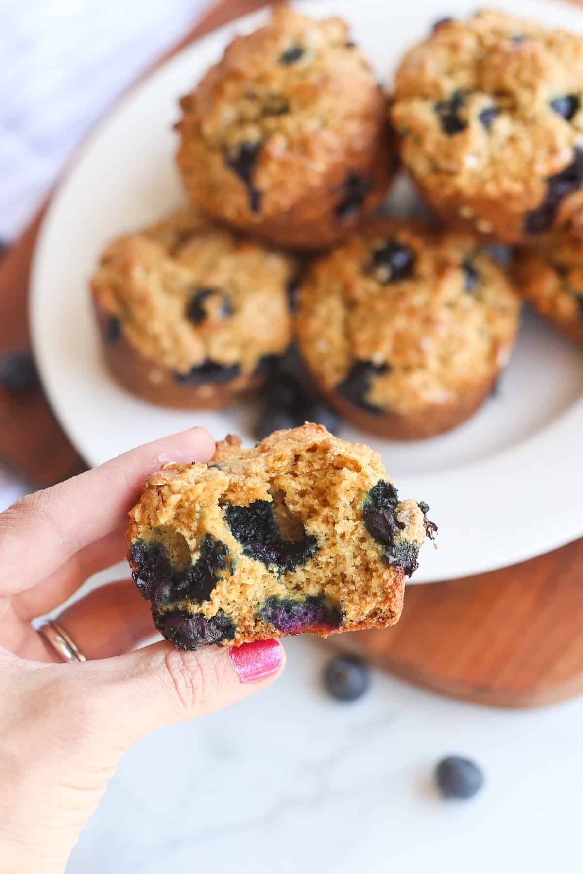 A hand holding a blueberry oatmeal muffin cut in half with a plate of the muffins in the background.