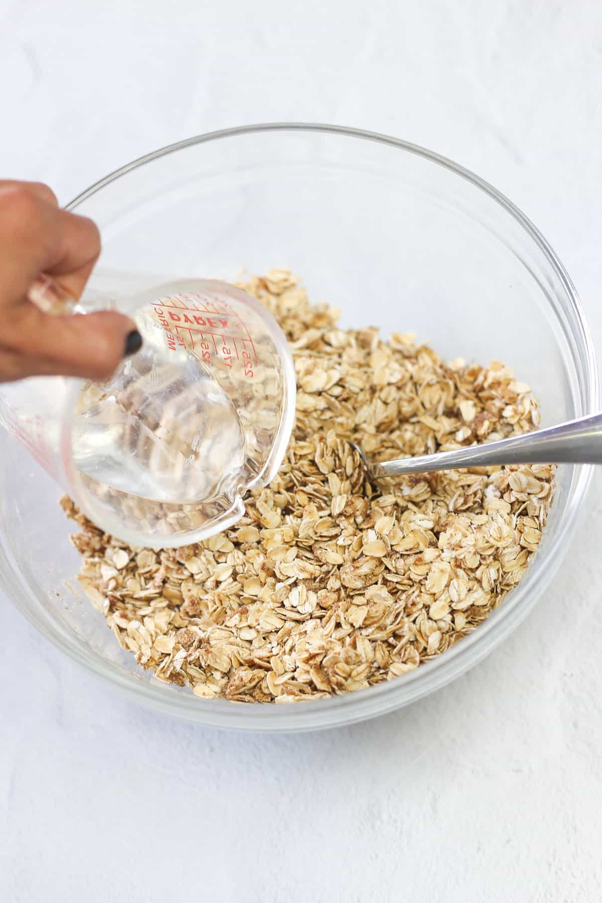 A hand pouring coconut oil into a mixing bowl of dry ingredients for baked oatmeal.