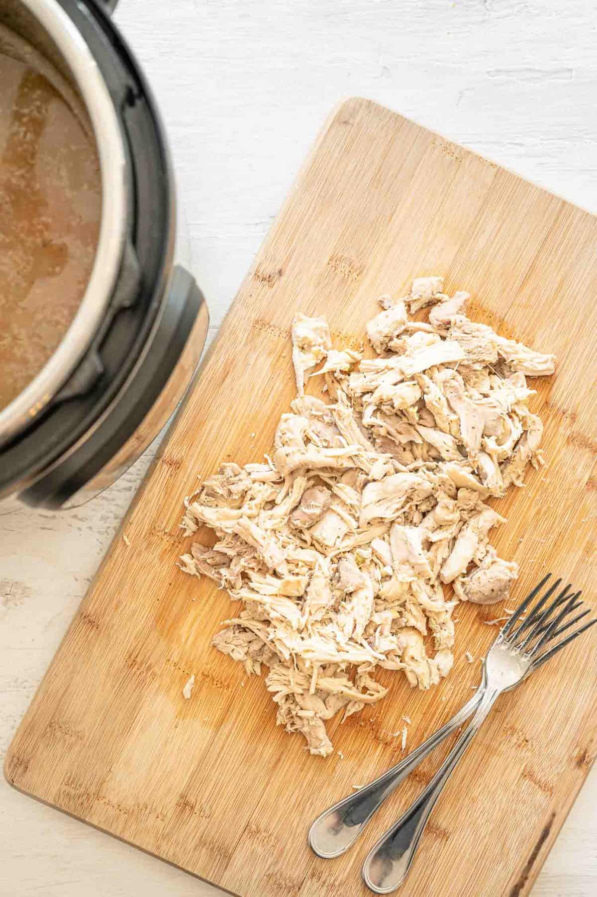 Shredded chicken on cutting board with two forks next to Instant Pot.