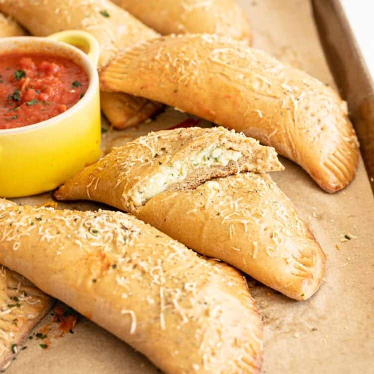 Calzones piled on baking sheet with one cut open and a small crock of pizza sauce.