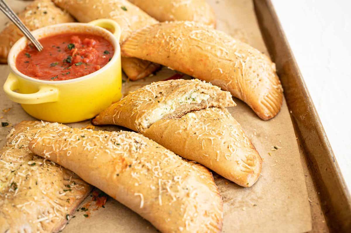 Calzones after baking piled on baking sheet with one cut in half and a small crock of pizza sauce.