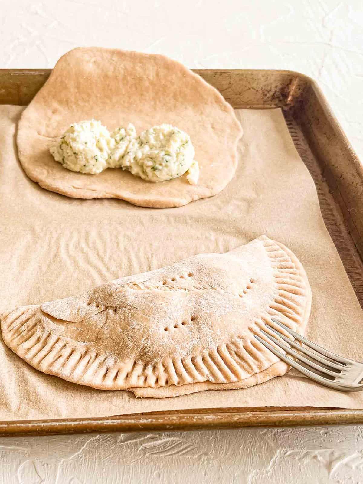 Calzone Dough on a baking sheet with scoops of the cheesy filling on them. One has been folded over and fork marks are being made along the edges to seal it.