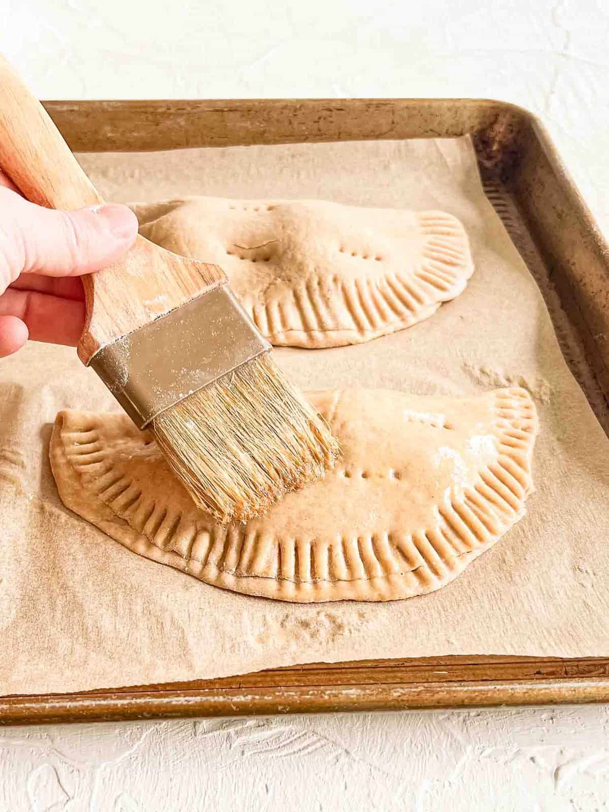 Two calzones on baking sheet getting an egg wash put on with a pastry brush.