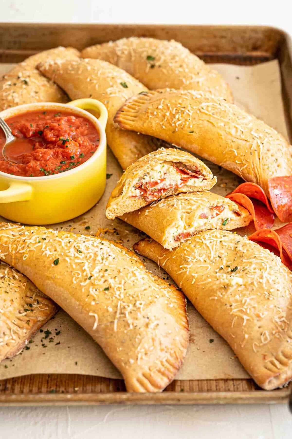 Pepperoni Calzones piled on baking sheet with one cut open to see the cheesy insides and pepperoni slices.