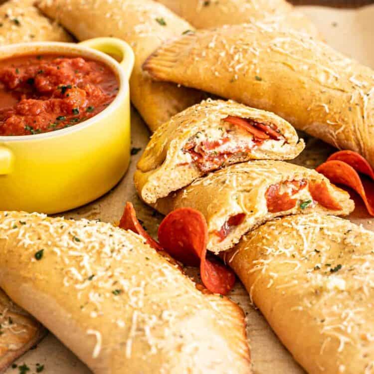 Pepperoni Calzones piled on baking sheet with one cut open to see the cheesy insides and pepperoni.