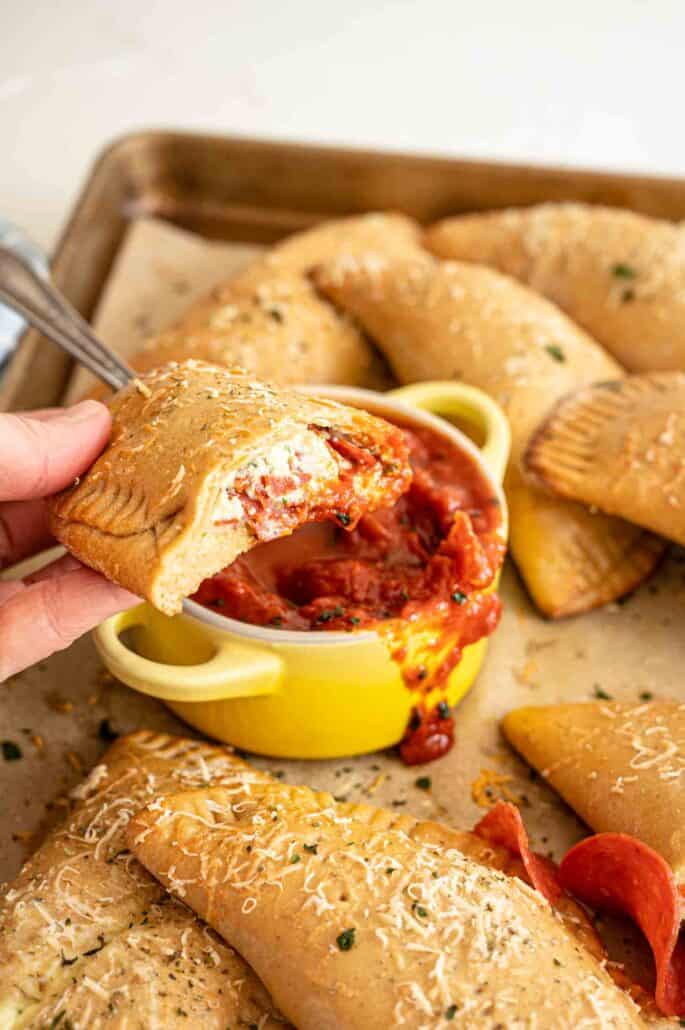 Pepperoni Calzones on baking sheet with one cut in half and being held above the others after having been dipped in marinara.