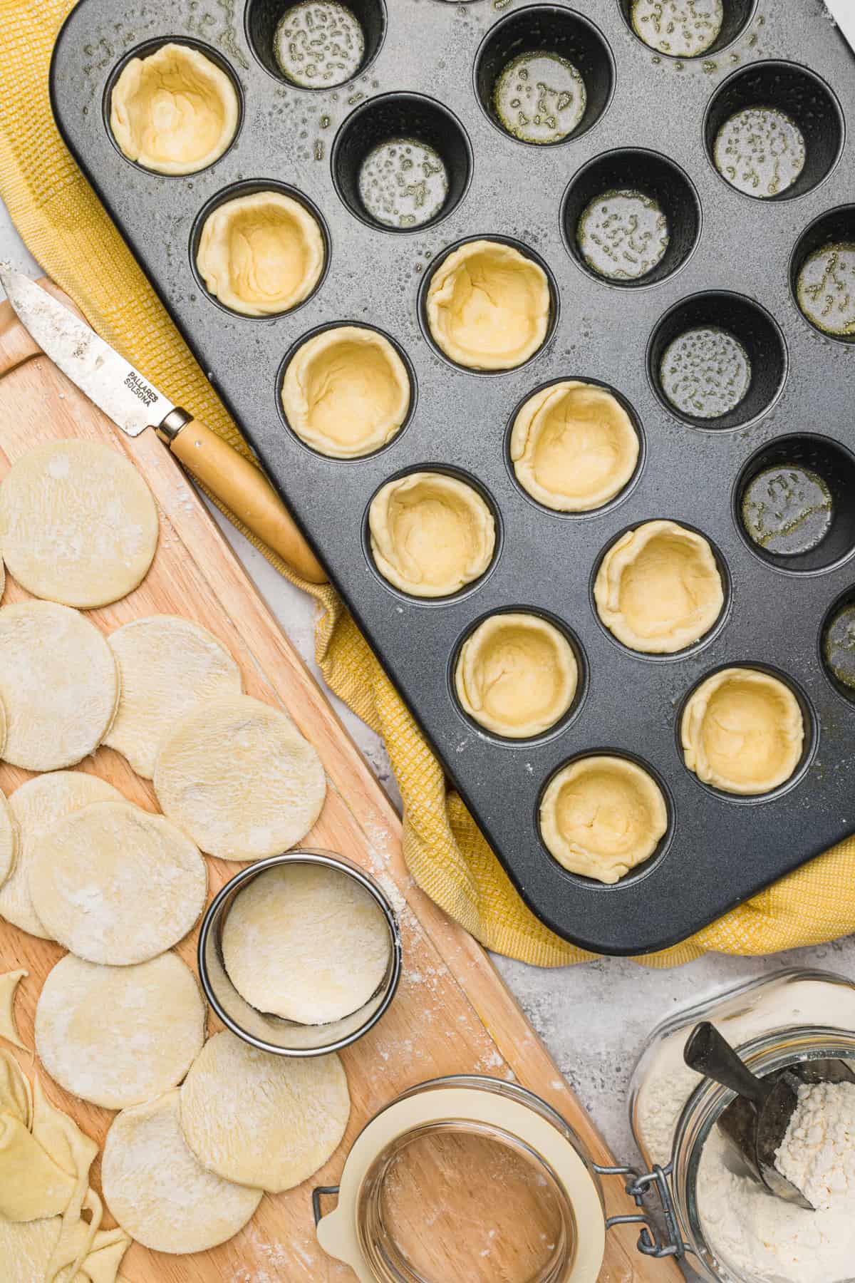 Wooden cutting board with pie crust circles on it next to a mini muffin pan that has been sprayed with oil. A few of the muffin tin cups already have pie crust circles pushed into them.