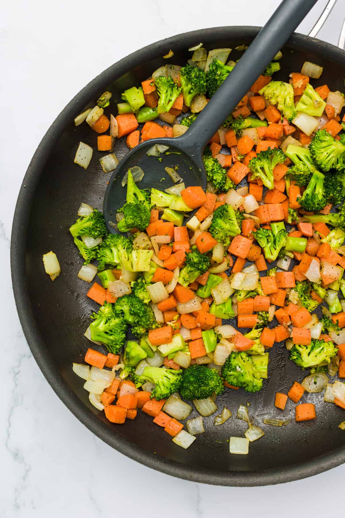 Chopped carrots, onions, and broccoli being sautéed in a pan.