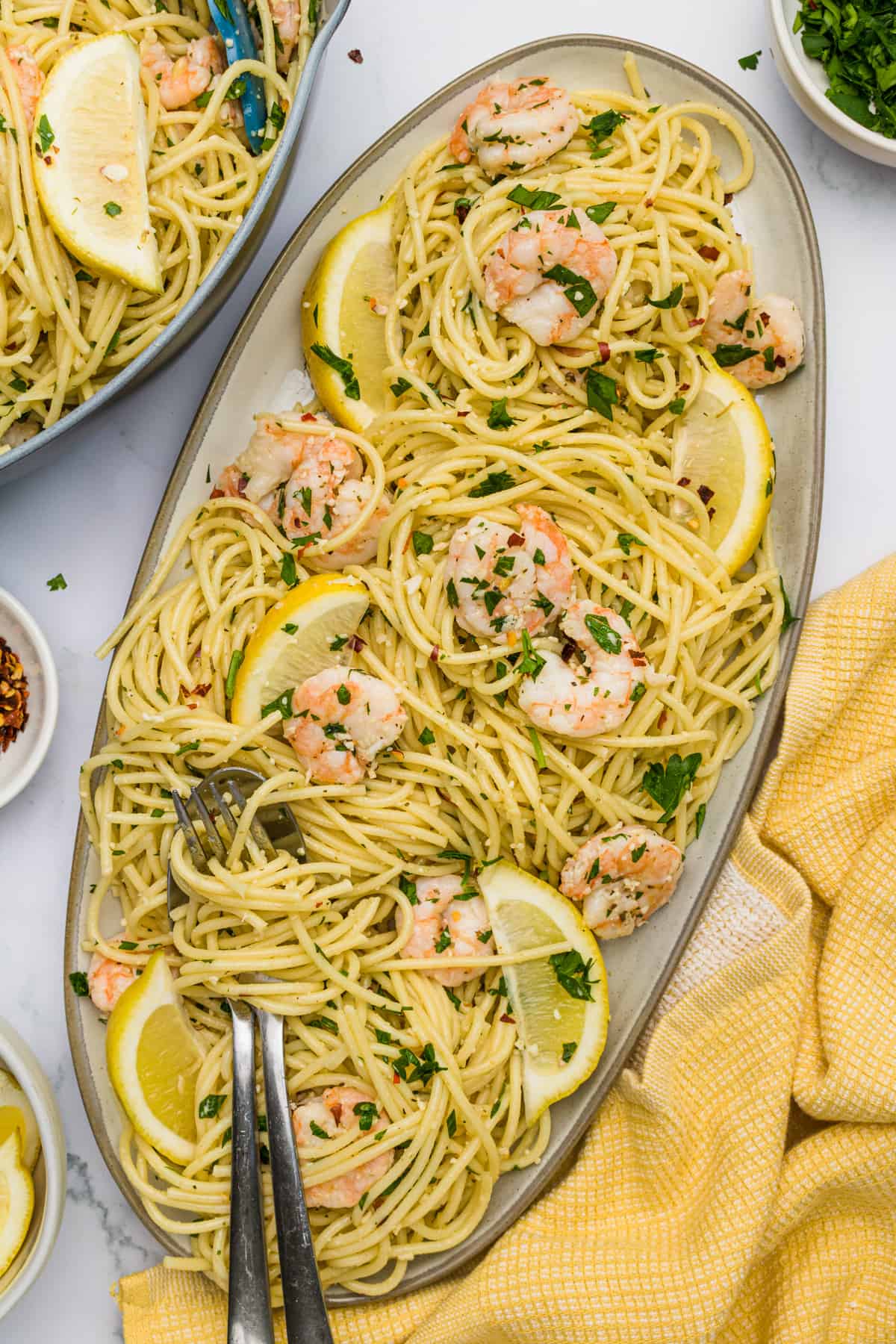 Overhead close up of Shrimp Scampi pasta in an oblong ceramic casserole dish with a metal fork and spoon twisted into the pasta sitting on a yellow dish towel.