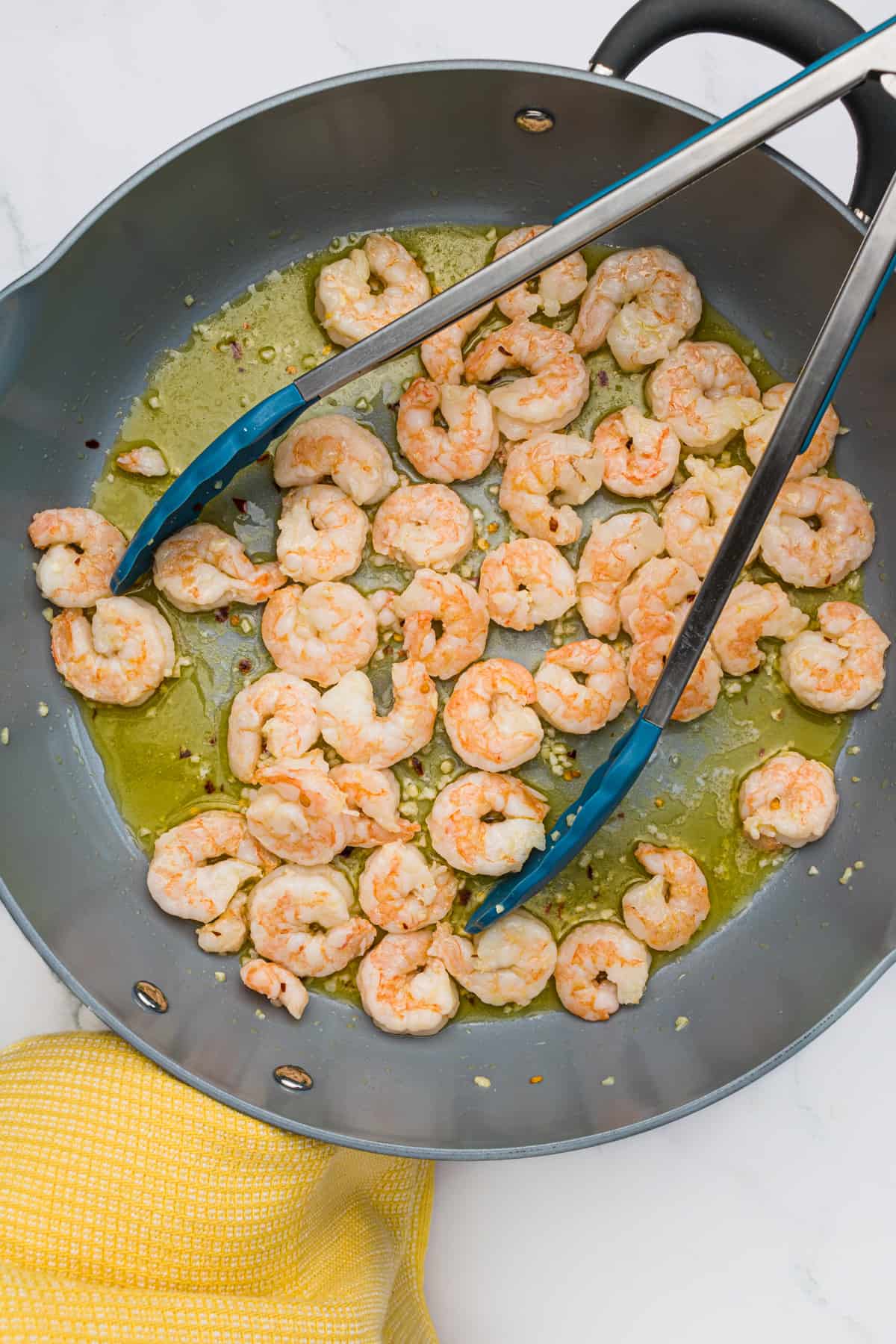 Shrimp cooking in oil and butter with minced garlic in a skillet with tongs.