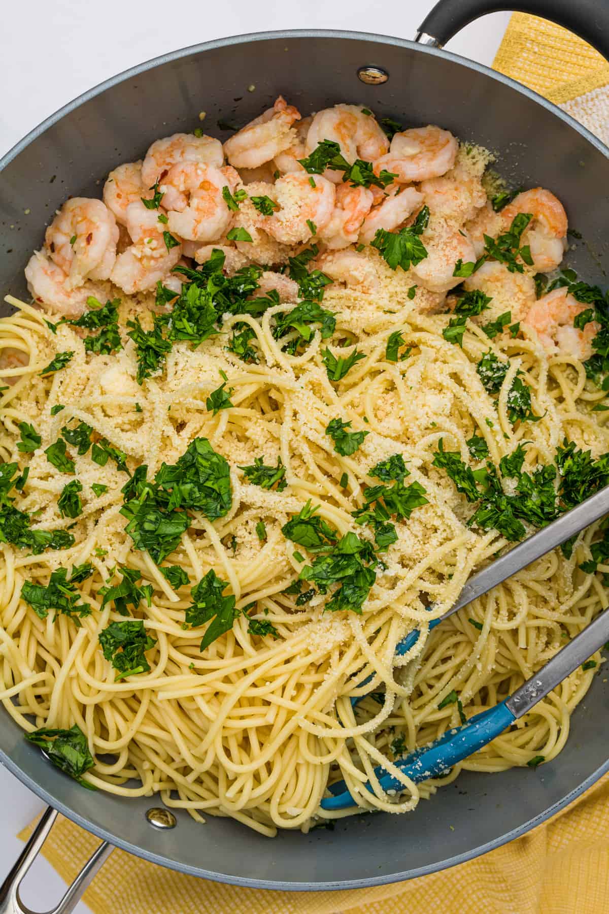 Metal skillet with spaghetti noodles, cooked shrimp, chopped fresh parsley and grated Parmesan cheese being mixed together with tongs.