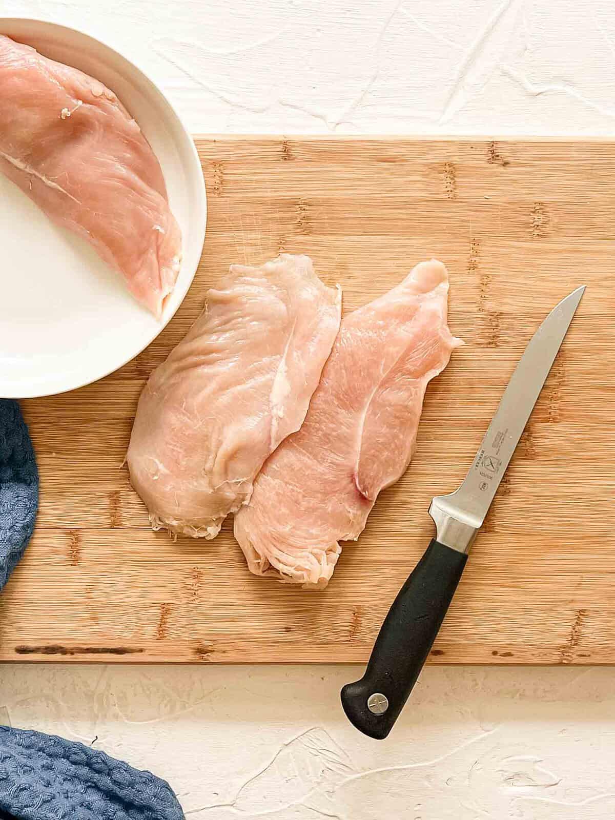Raw chicken breast on cutting board with knife having been sliced in half lengthwise.