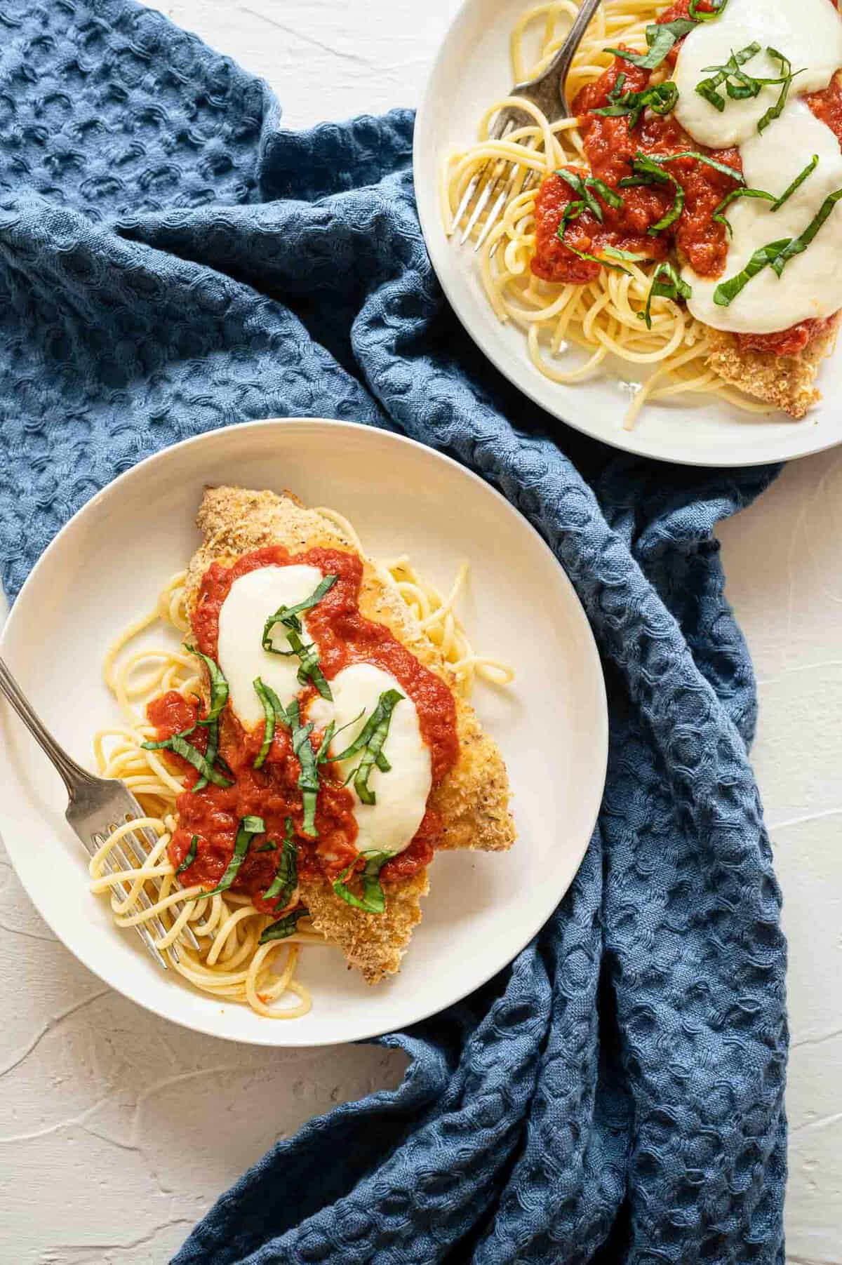 Two plates of baked chicken parmesan on top of whole wheat noodles.