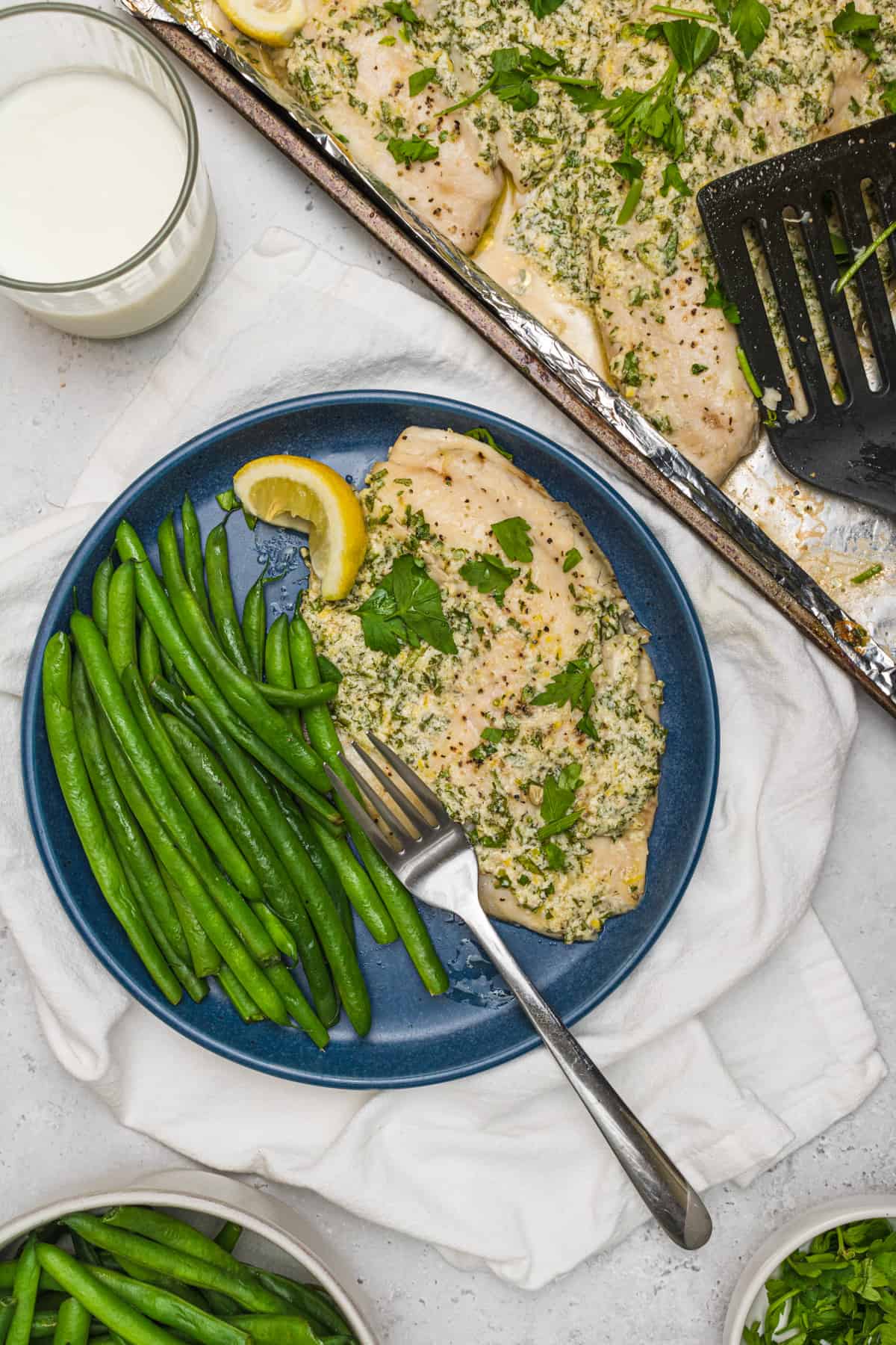 Parmesan Broiled Tilapia on a dark blue plate with long green beans and a fork on it. There is a pan of cooked Parmesan Tilapia to the top right and a glass of milk to the top left.