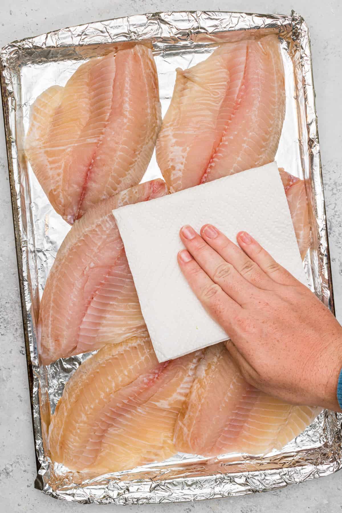 Raw tilapia fillets on baking sheet lined with foil. A hand is using a paper towel to pat the fillets dry.