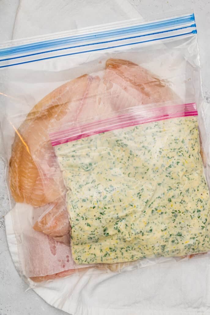 Raw tilapia fillets and a small bag with the mixed together coating for Parmesan Broiled Tilapia inside a gallon-size freezer bag.