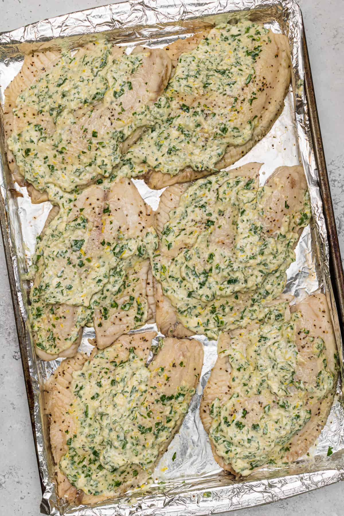 Raw tilapia fillets on baking sheet lined with foil with the lemon parmesan mixture on top of each one.