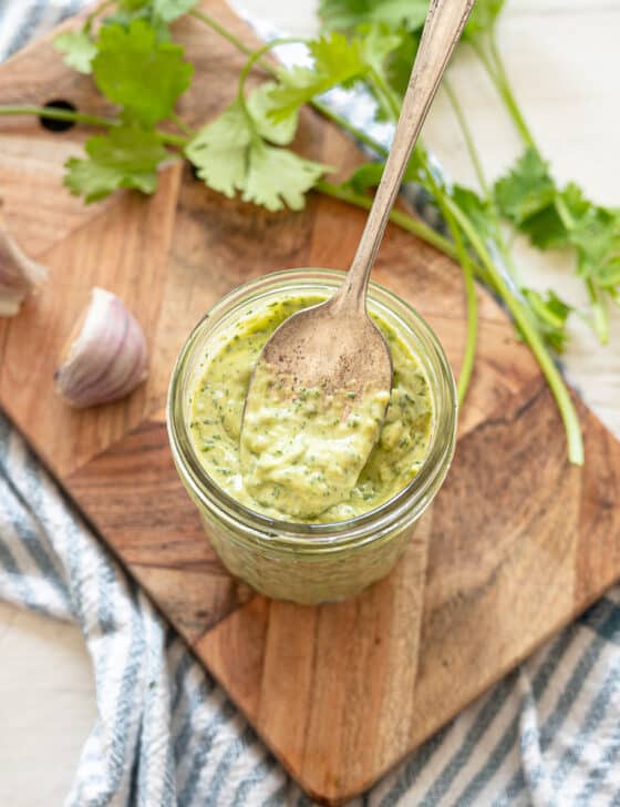 Cilantro Lime Aioli in a small mason jar with a spoon dipped in, sitting on a wooden cutting board with lime halves, garlic cloves, and fresh cilantro around it.