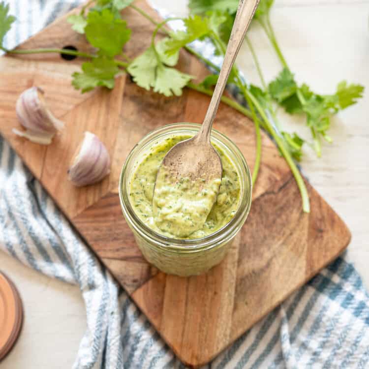 Cilantro Lime Aioli in a small mason jar with a spoon dipped in, sitting on a wooden cutting board with lime halves, garlic cloves, and fresh cilantro around it.