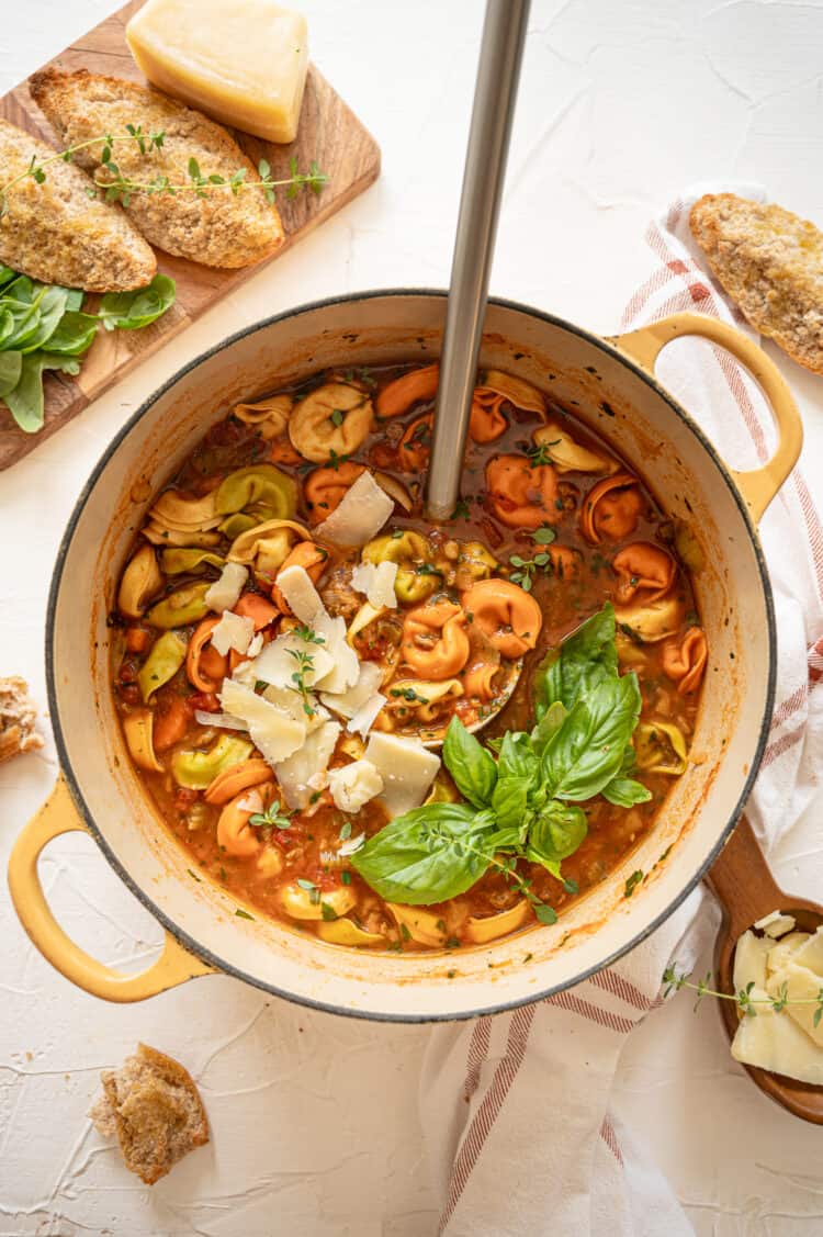 Italian sausage and tortellini soup fully cooked in a dutch oven with Parmesan cheese shavings and fresh basil on top.