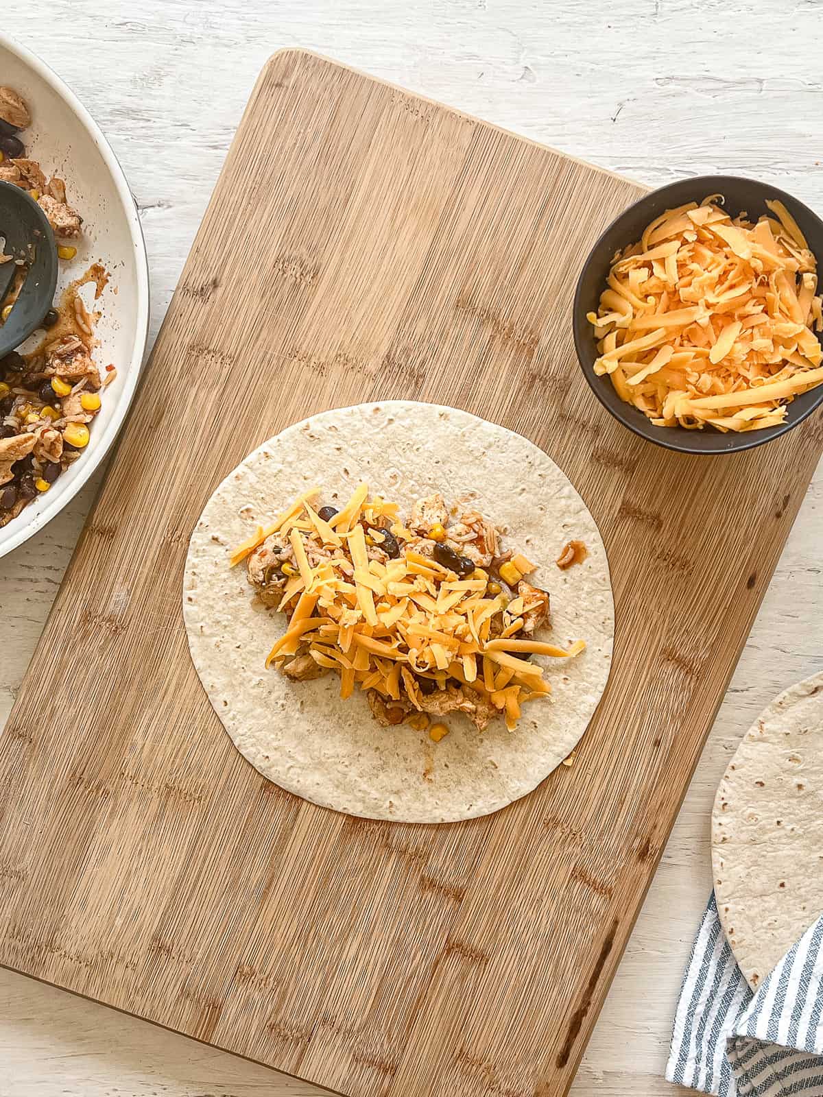 Chicken Burrito filling topped with shredded cheese laying on an open tortilla which is laying on a wooden cutting board.