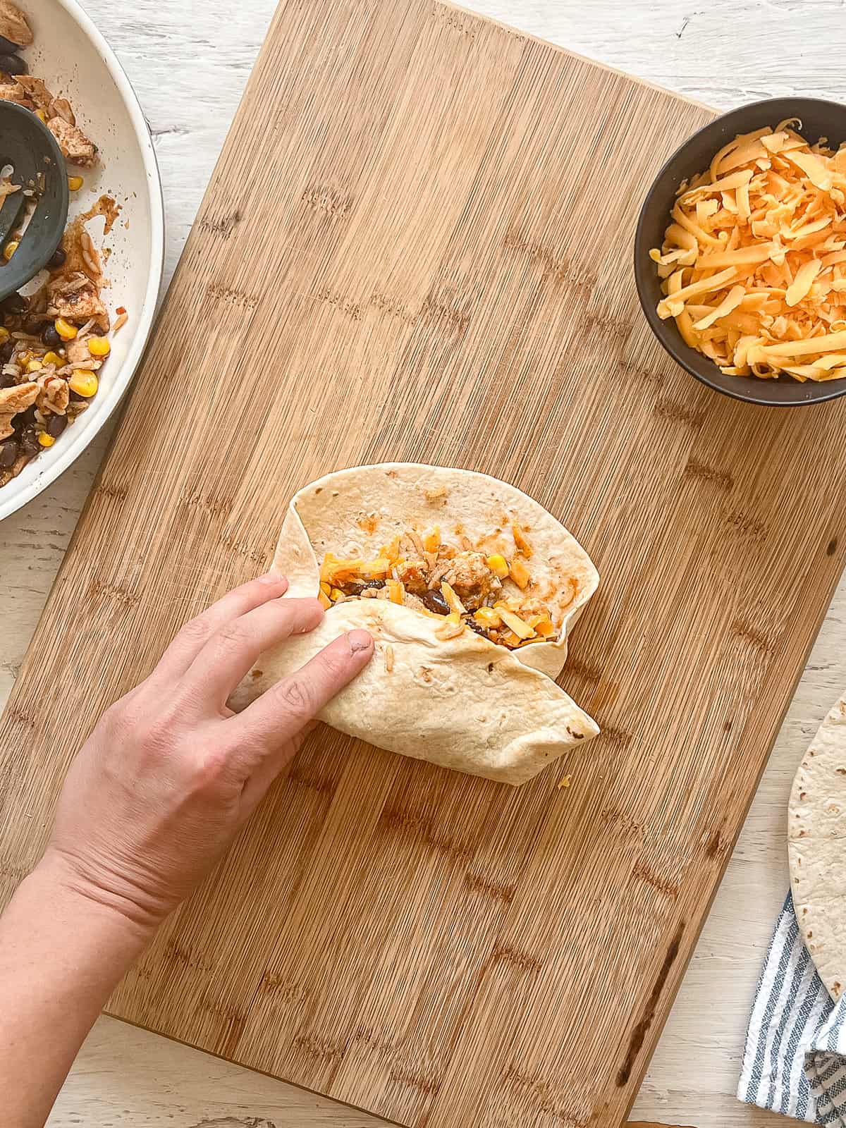 One hand is holding a partially wrapped chicken burrito on a wooden cutting board. Two sides of the tortilla have been folded in and the bottom has been folded up.
