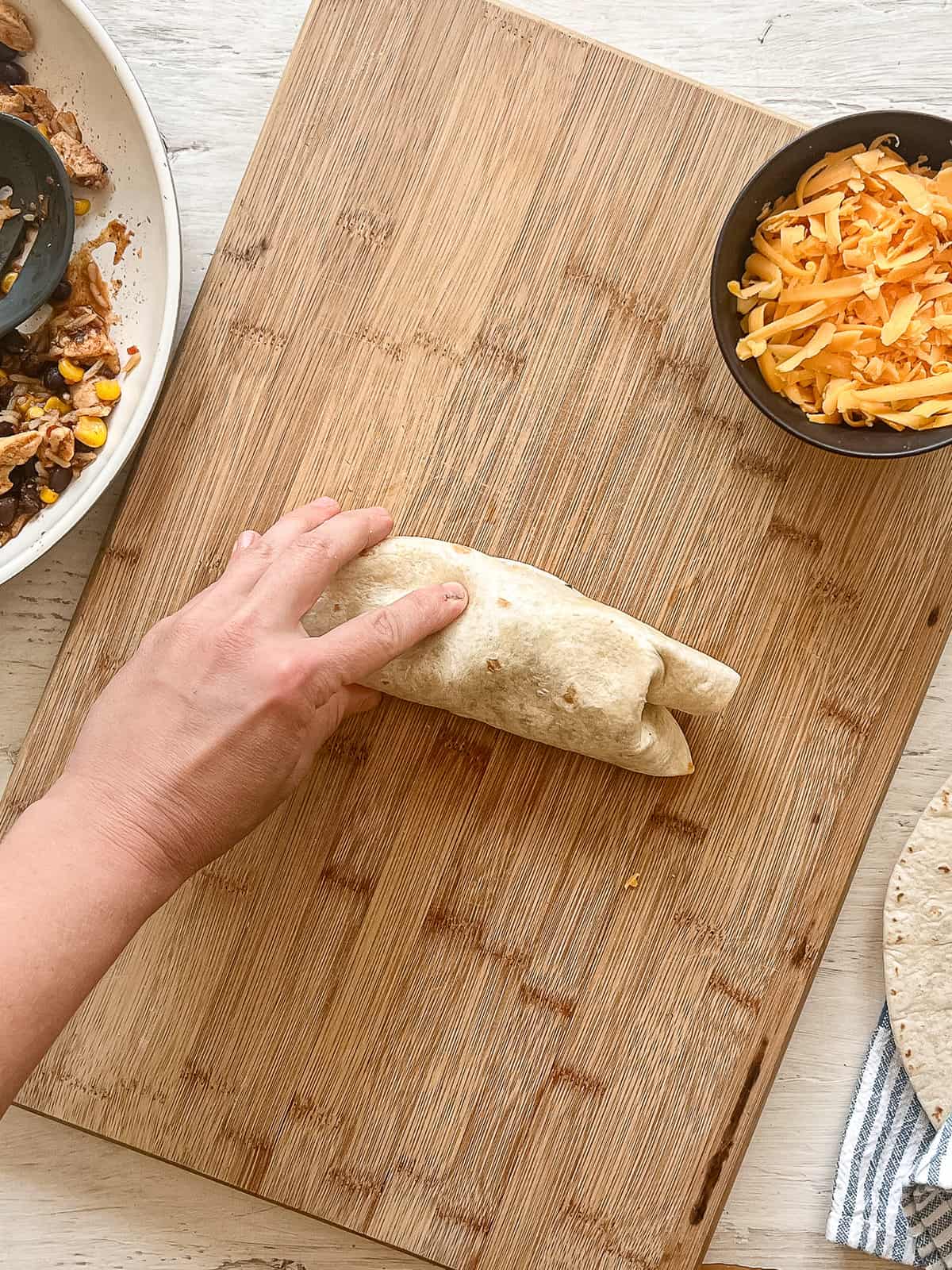 One hand is holding together a wrapped tortilla with chicken burrito ingredients inside on a wooden cutting board.