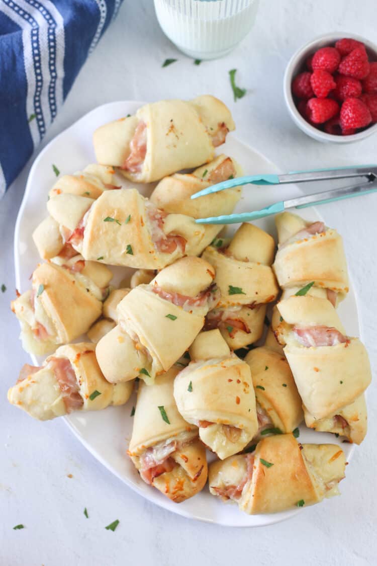 Ham and cheese croissants on a white platter with tongs.