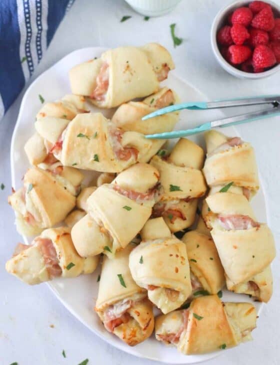 Ham and cheese croissants on a white platter with tongs.