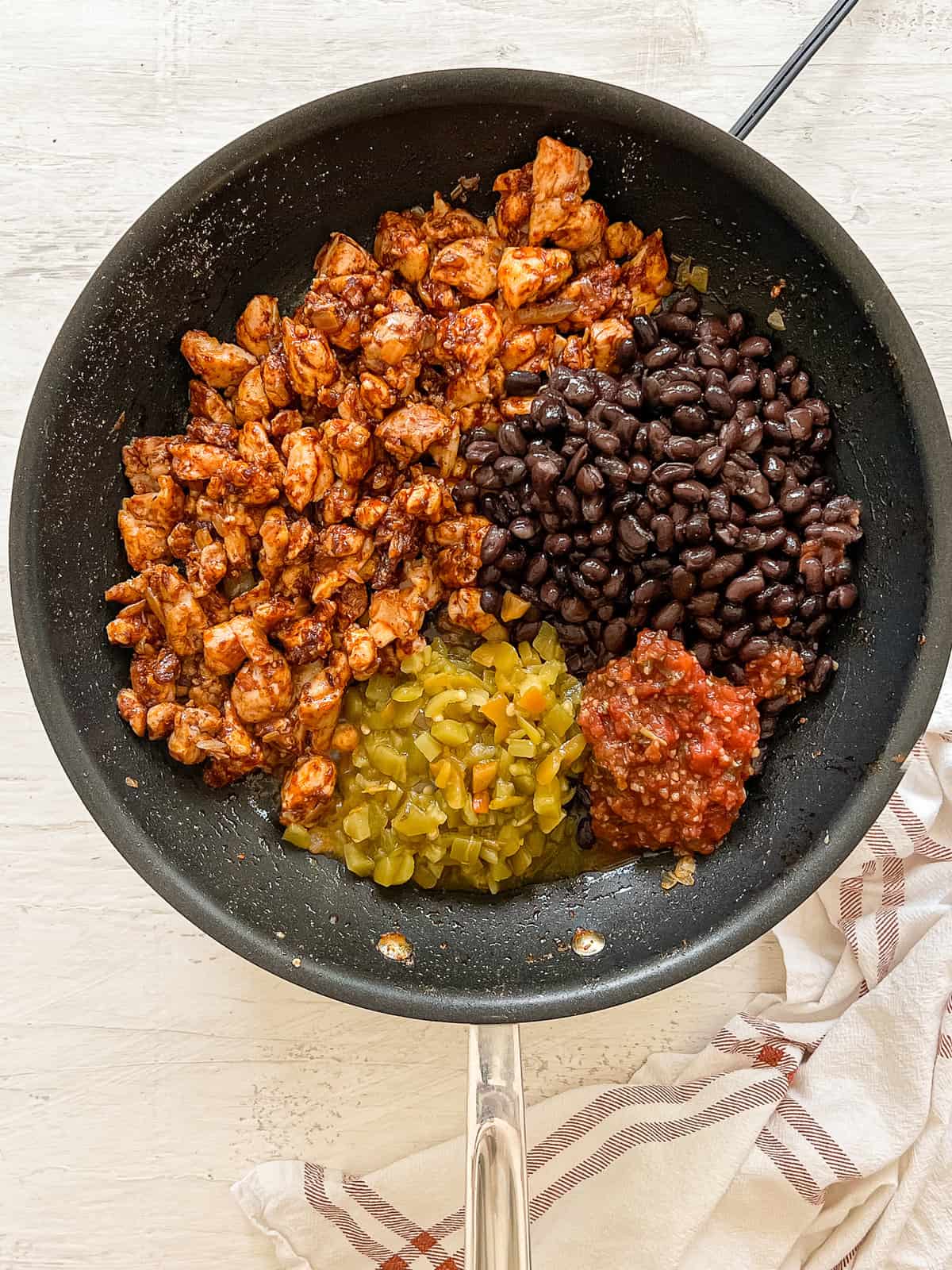 Cheesy Chicken Enchilada filling ingredients in piles in a skillet: cooked taco-seasoned chicken, green chiles, black beans, and salsa.