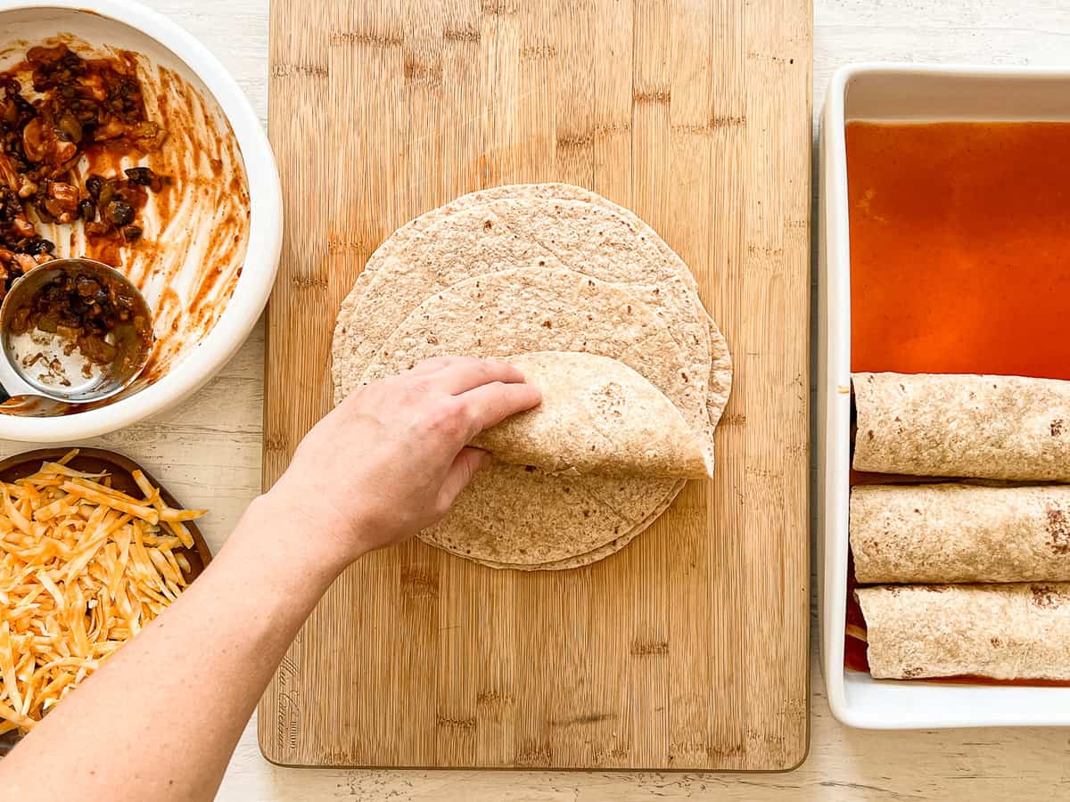 A hand rolling up a tortilla with chicken enchilada filling and shredded cheddar cheese in it with a casserole dish of rolled up tortillas and red enchilada sauce next to it.
