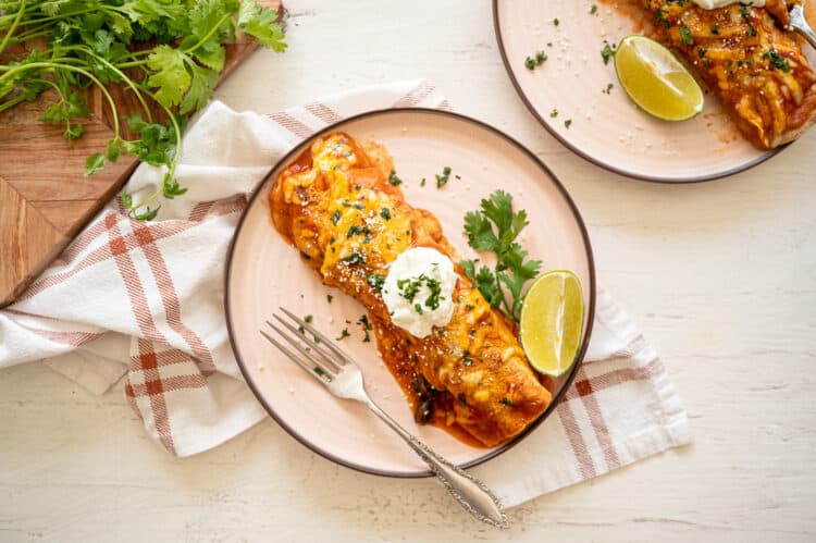 Cheesy Chicken Enchiladas served on two plates with sour cream on top and lime wedges and fresh cilantro on the side.