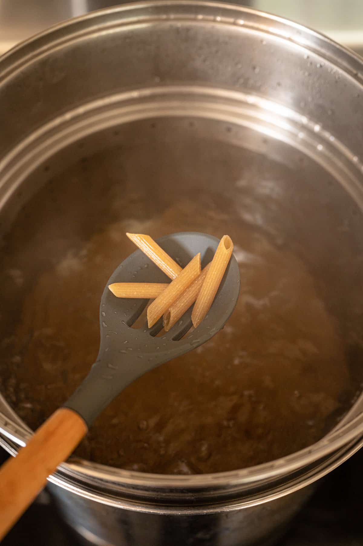 Slotted spoon with penne pasta on it above a pot of boiling penne.