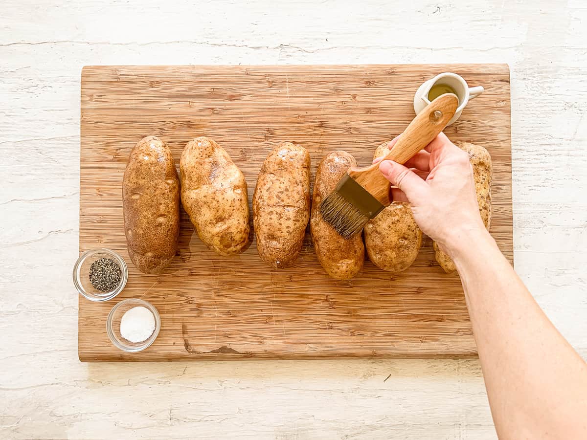 Six large baking potatoes sitting on a wooden cutting board and being brushed with oil with a pastry brush.